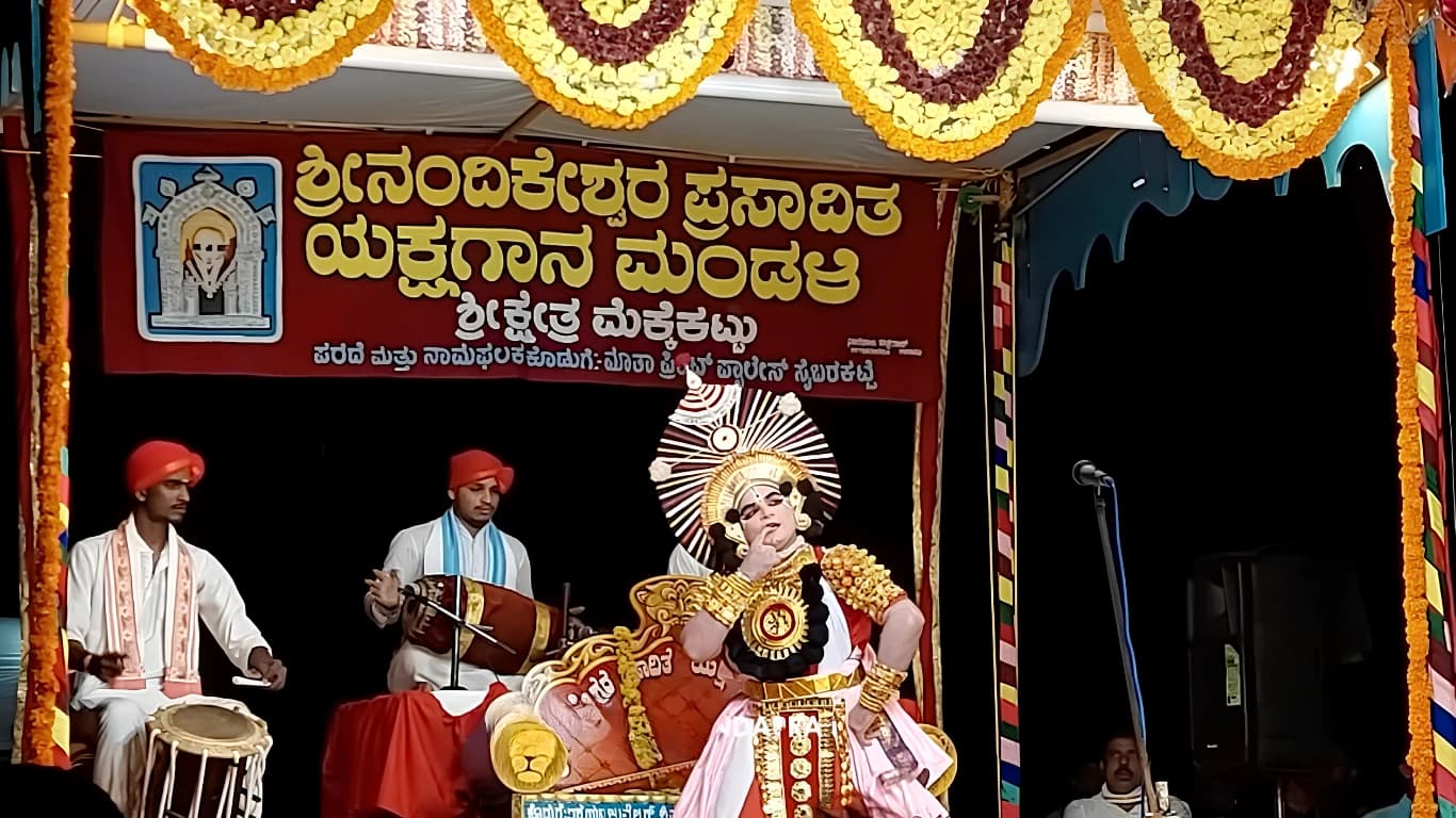 Yakshagana performer in painted make-up, gold ornaments and a large ornate headdress on stage, with two drummers in white dress and red headcloths seated beside him, below the Mandali's red banner and rows of marigold garlands